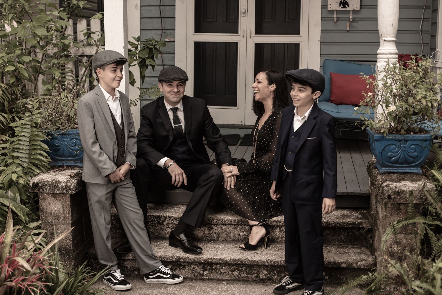 Four people in vintage-style clothing and caps sit together on stone steps of a historic house.