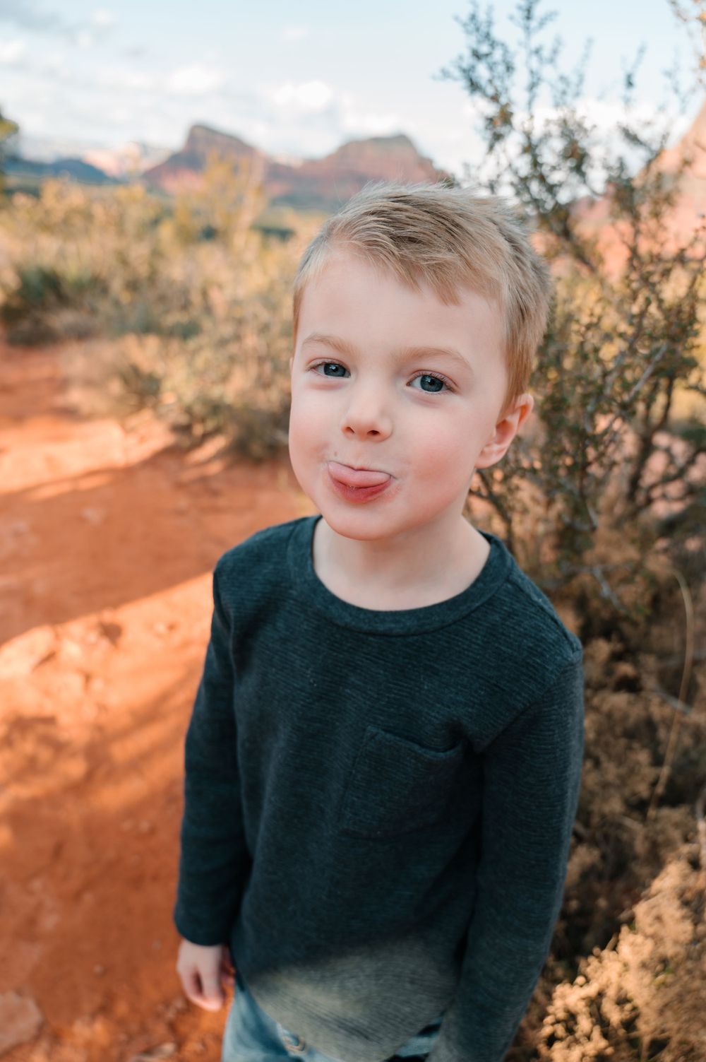 A child in a black sweater smiles while sitting on red rocks in a desert landscape.