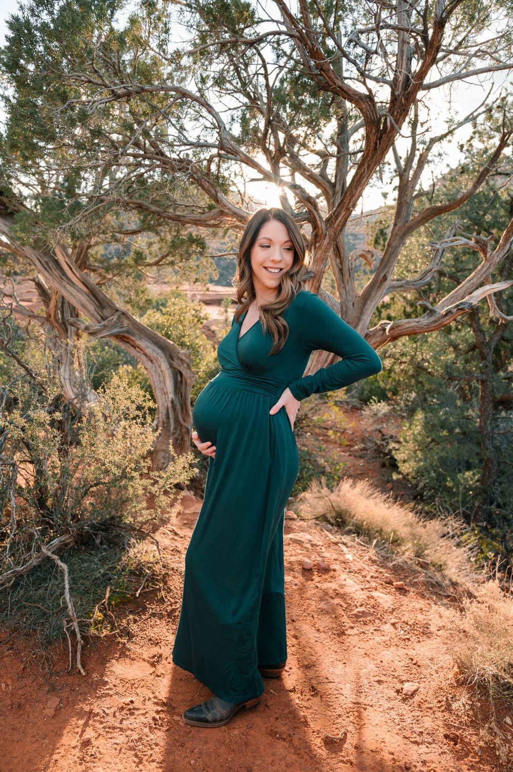 A woman in a dark green dress poses playfully on red rocks surrounded by desert vegetation.