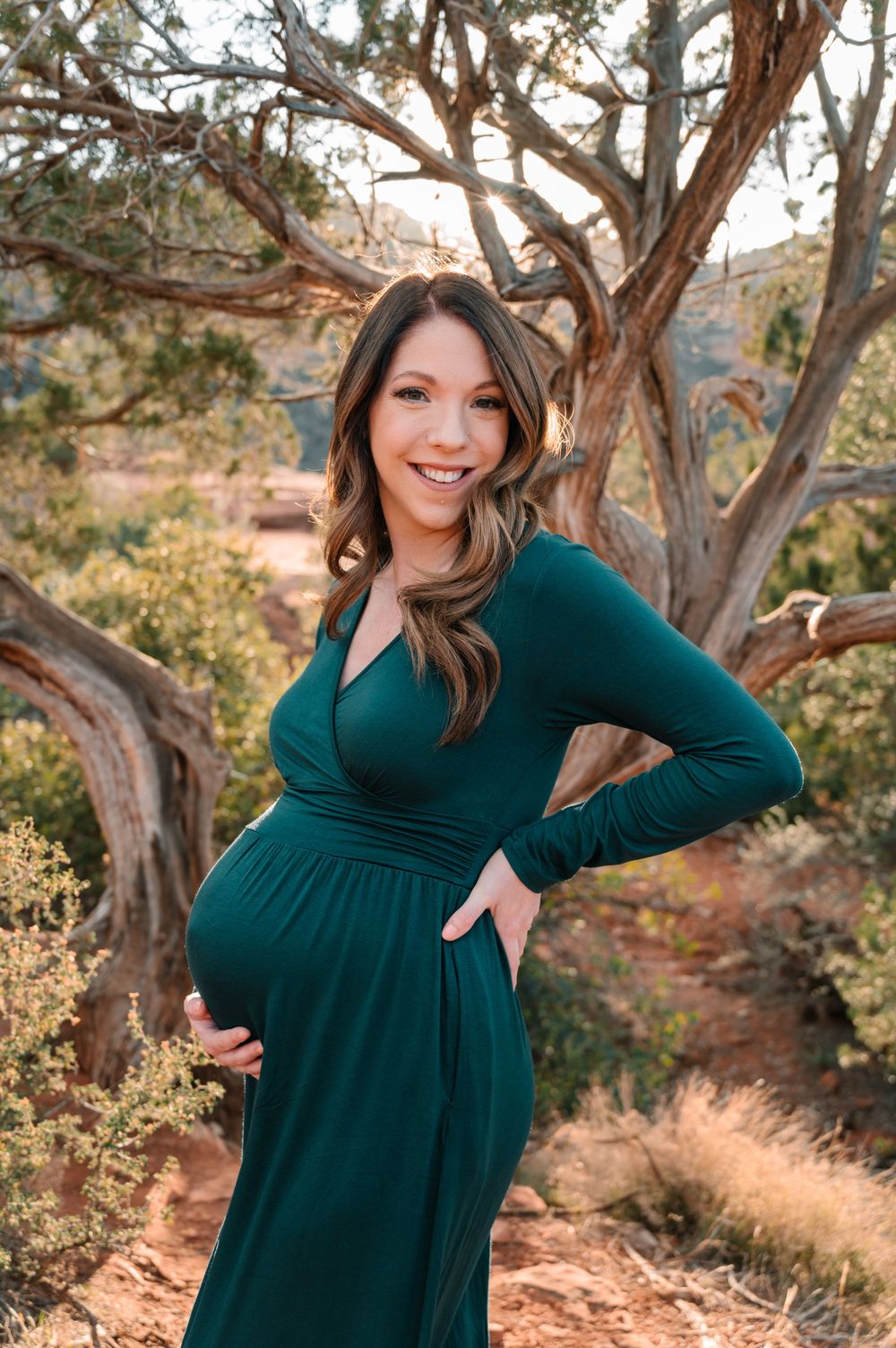 A pregnant woman in emerald dress poses joyfully against a backdrop of desert scenery.