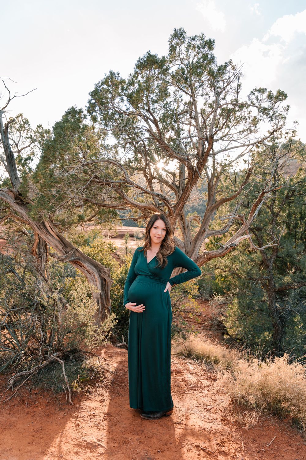 A woman in green maternity dress stands among juniper trees on red desert soil.