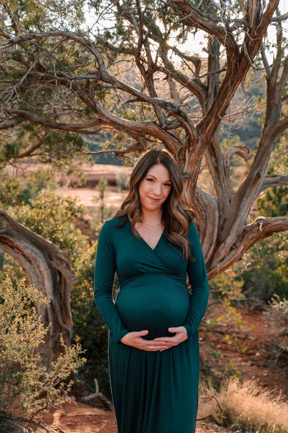 Woman in emerald dress poses thoughtfully in a desert landscape with twisted juniper trees at sunset.