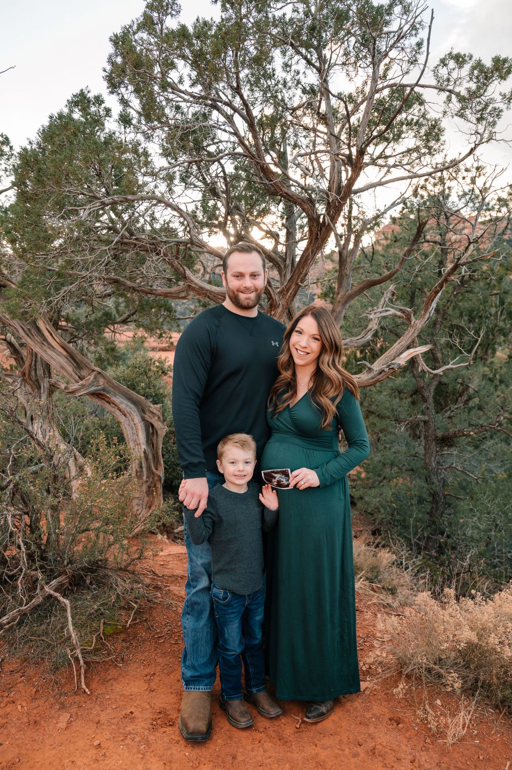 Family of three poses together among desert juniper trees at sunset.