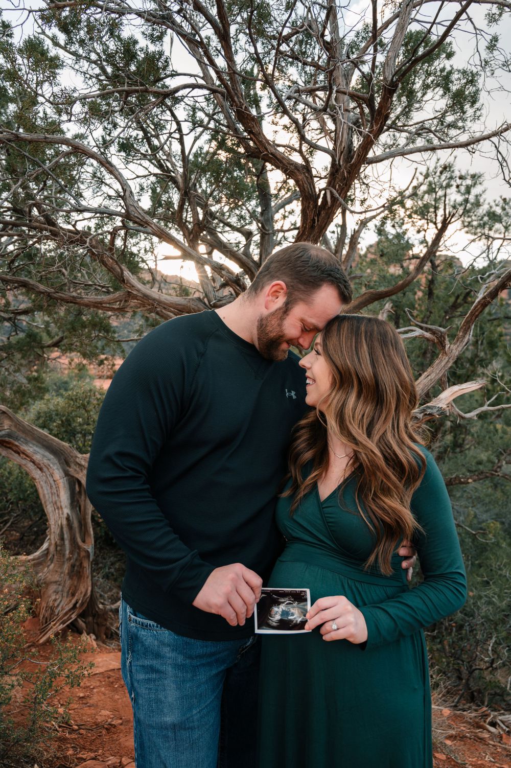 Couple embraces while looking at ultrasound photo among desert juniper trees.