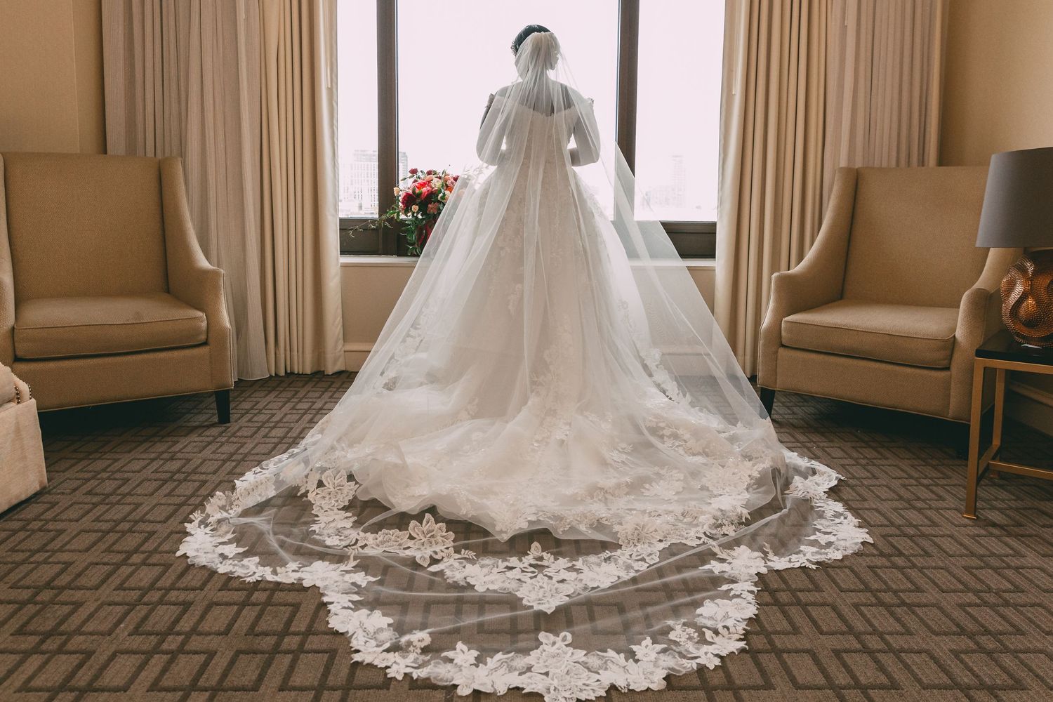 Stunning lace wedding gown with cathedral veil displayed from behind near sunlit windows in a hotel room.