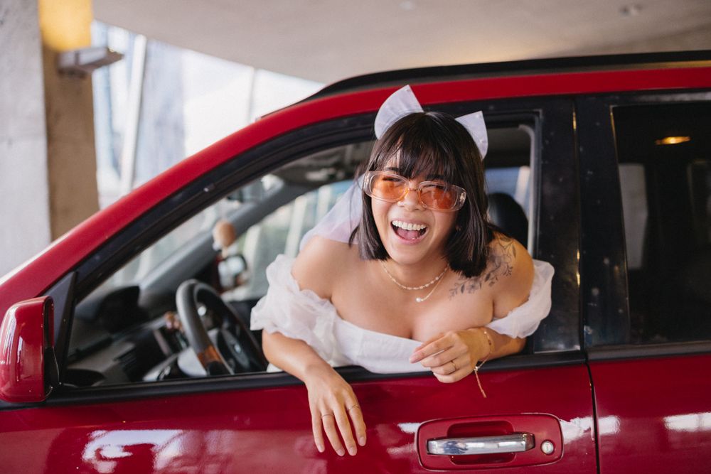 Whimsical poses next to a red car in a parking garage wearing a white dress.