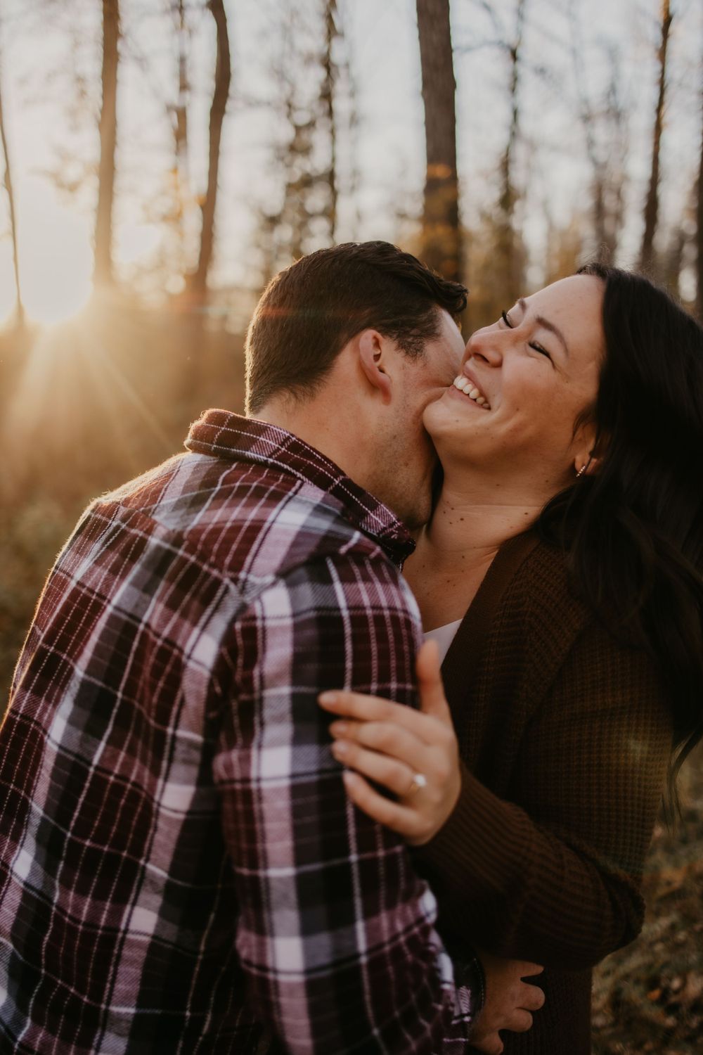A romantic couple shares an intimate laughing moment in the woods during golden hour.
