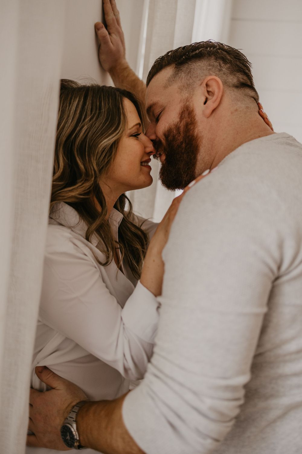 Romantic couple sharing an intimate moment against a white wall in matching cream sweaters.