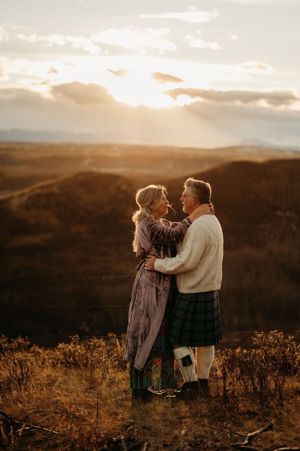 A couple embraces on a hilltop at sunset with one person wearing a traditional Scottish kilt.