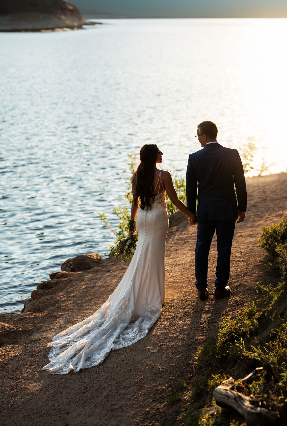 A couple walks along a waterfront path at sunset with flowing wedding dress trailing behind.