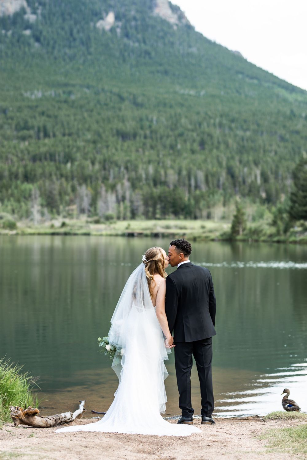 A couple shares an intimate moment by a serene mountain lake with evergreen forests in the background.