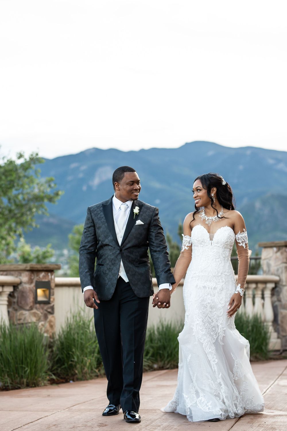 A couple in wedding attire walks together on a terrace with mountain views in the background.