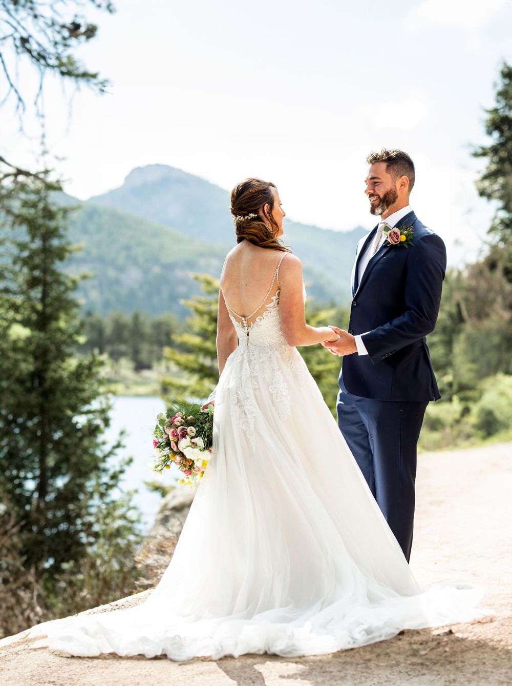 A couple in wedding attire stands together on a mountain path with evergreen trees and peaks in the distance.