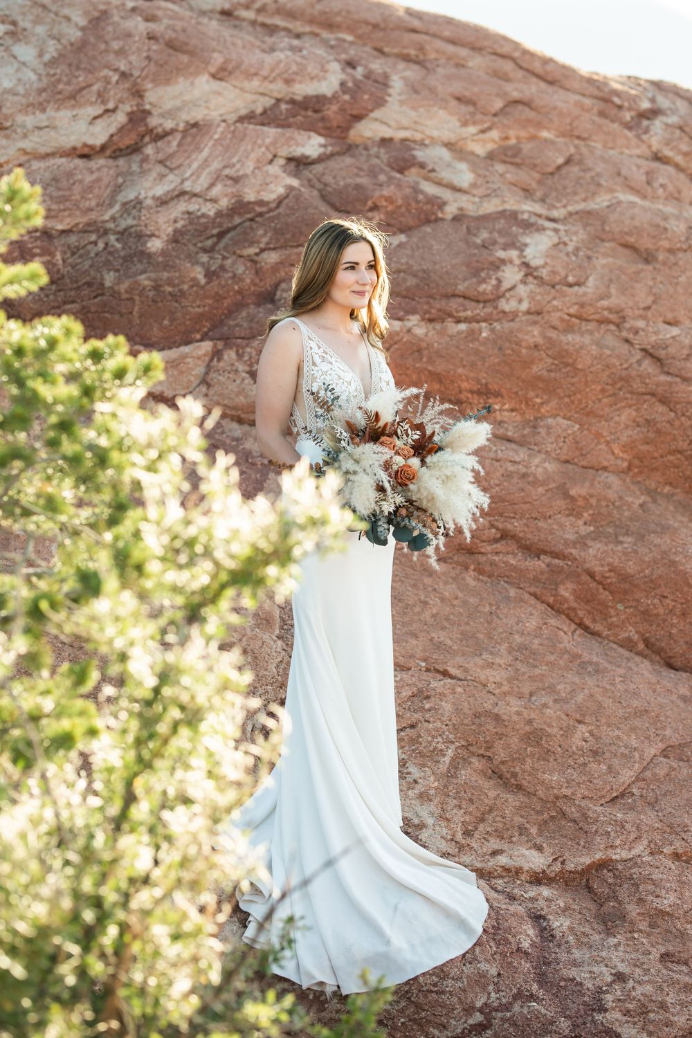 A bride in a white lace gown holds a bouquet while posing against red rock formations in golden sunlight.