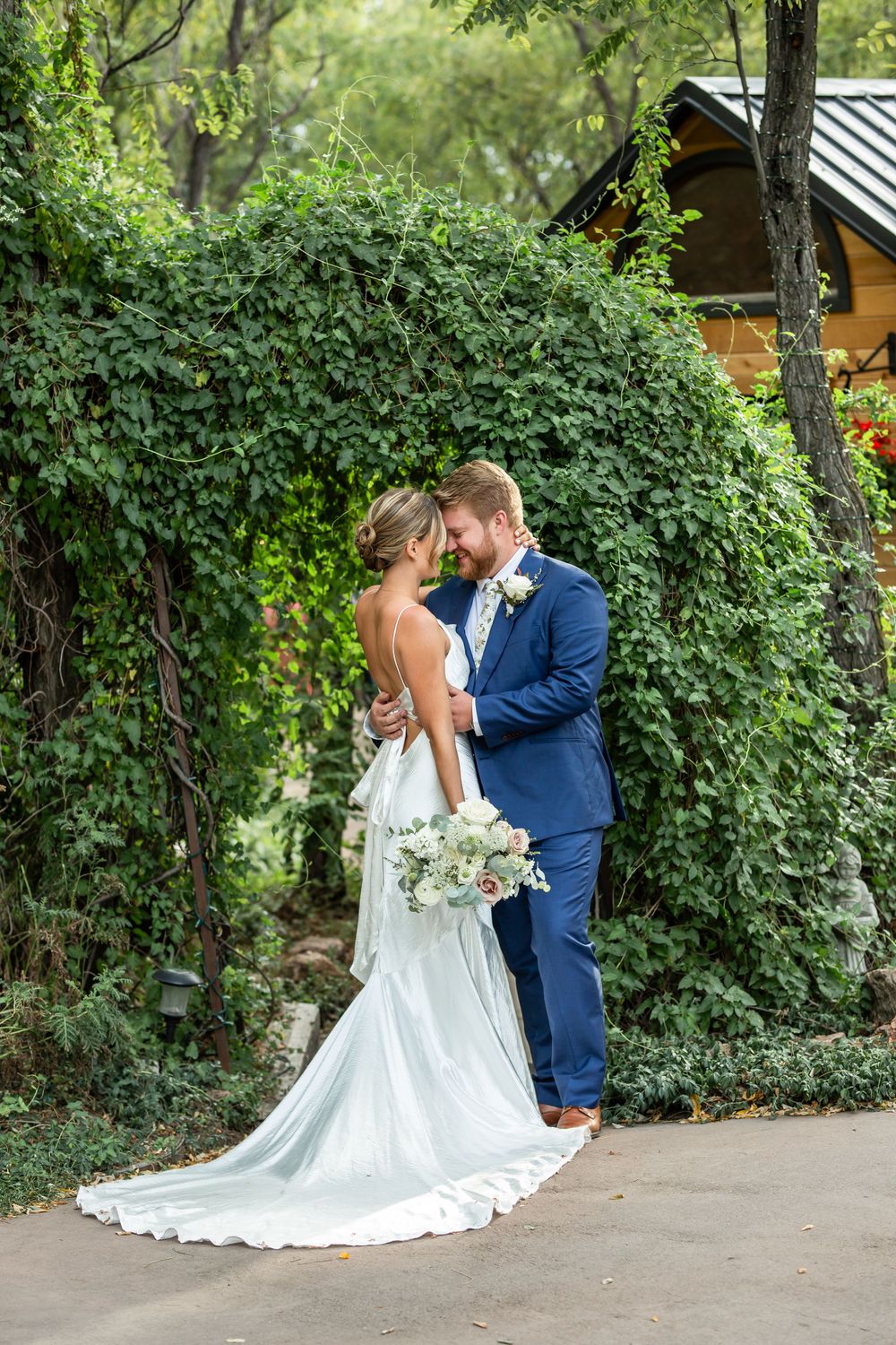 A wedding couple embraces under an archway of green foliage in a garden setting.