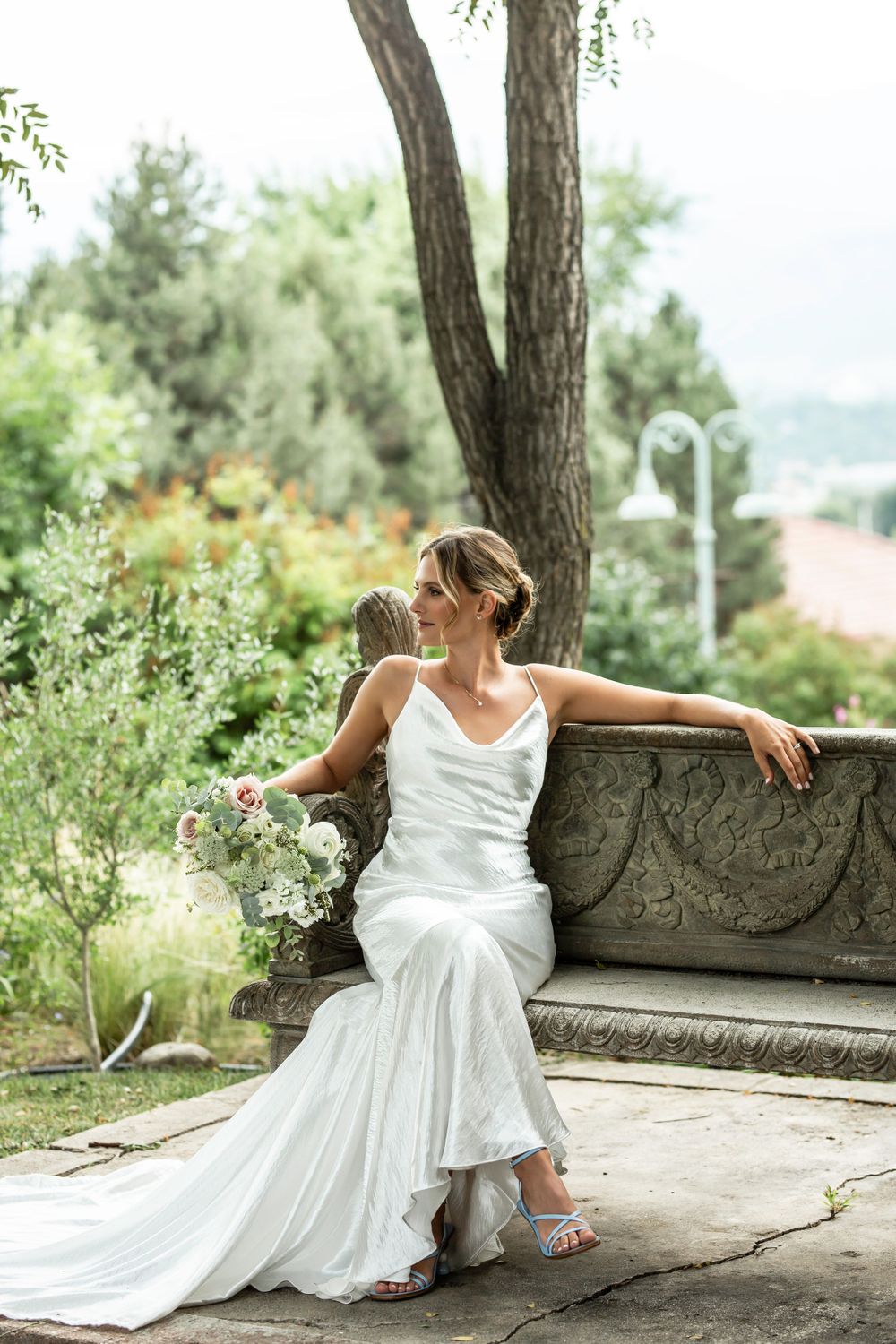 A bride in an elegant white dress poses gracefully on a stone bench in a garden setting.