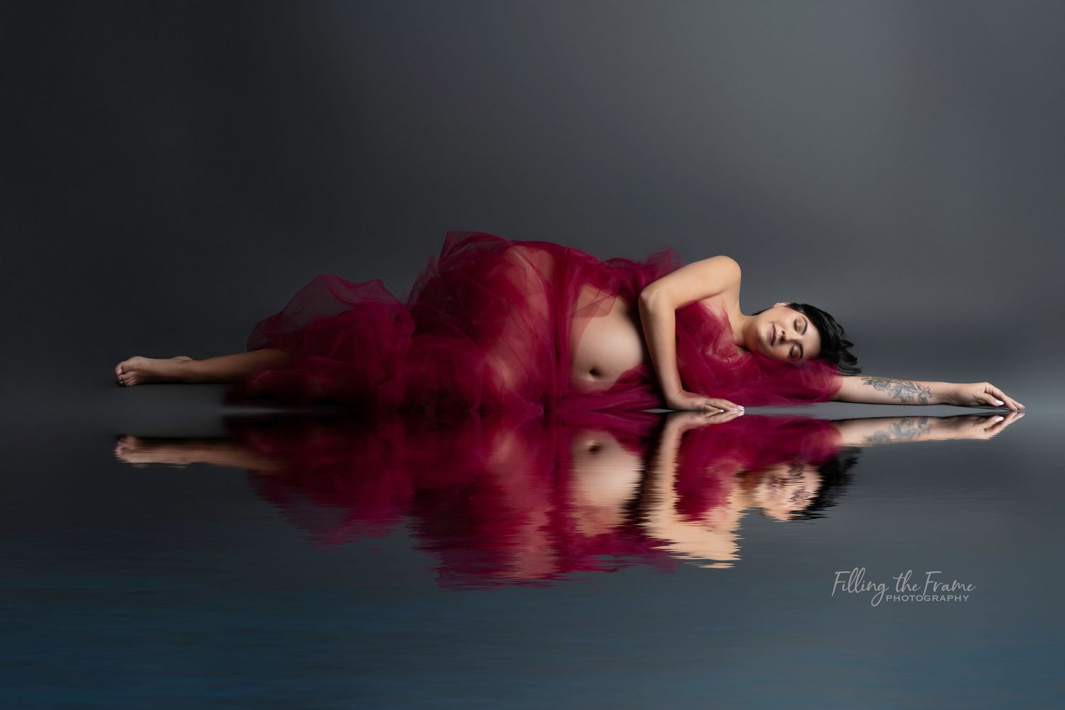 Woman in flowing burgundy dress poses gracefully on dark floor in artistic photo series.