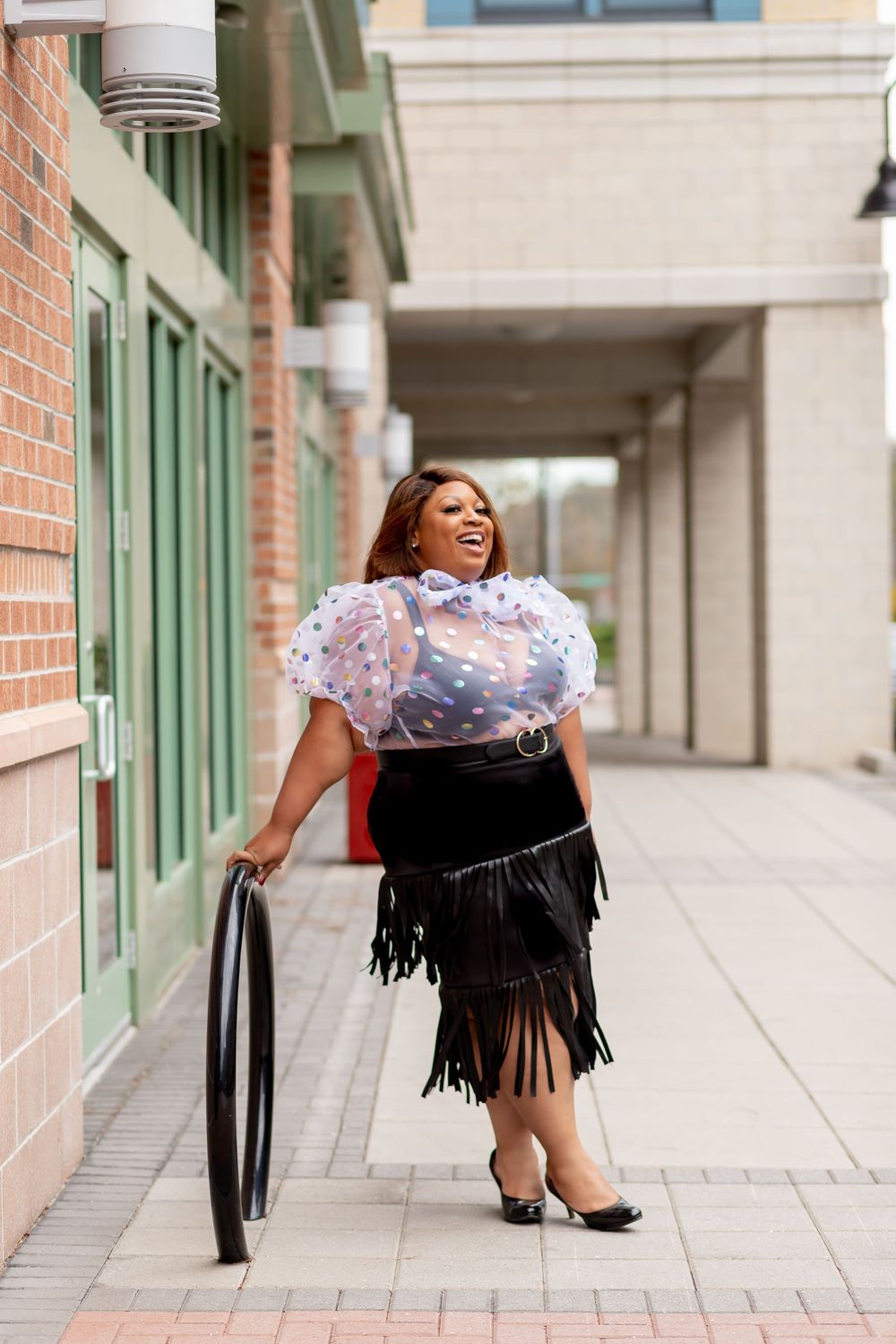 Plus-size woman in sheer polka dot top and black fringe skirt posing outdoors near brick building.
