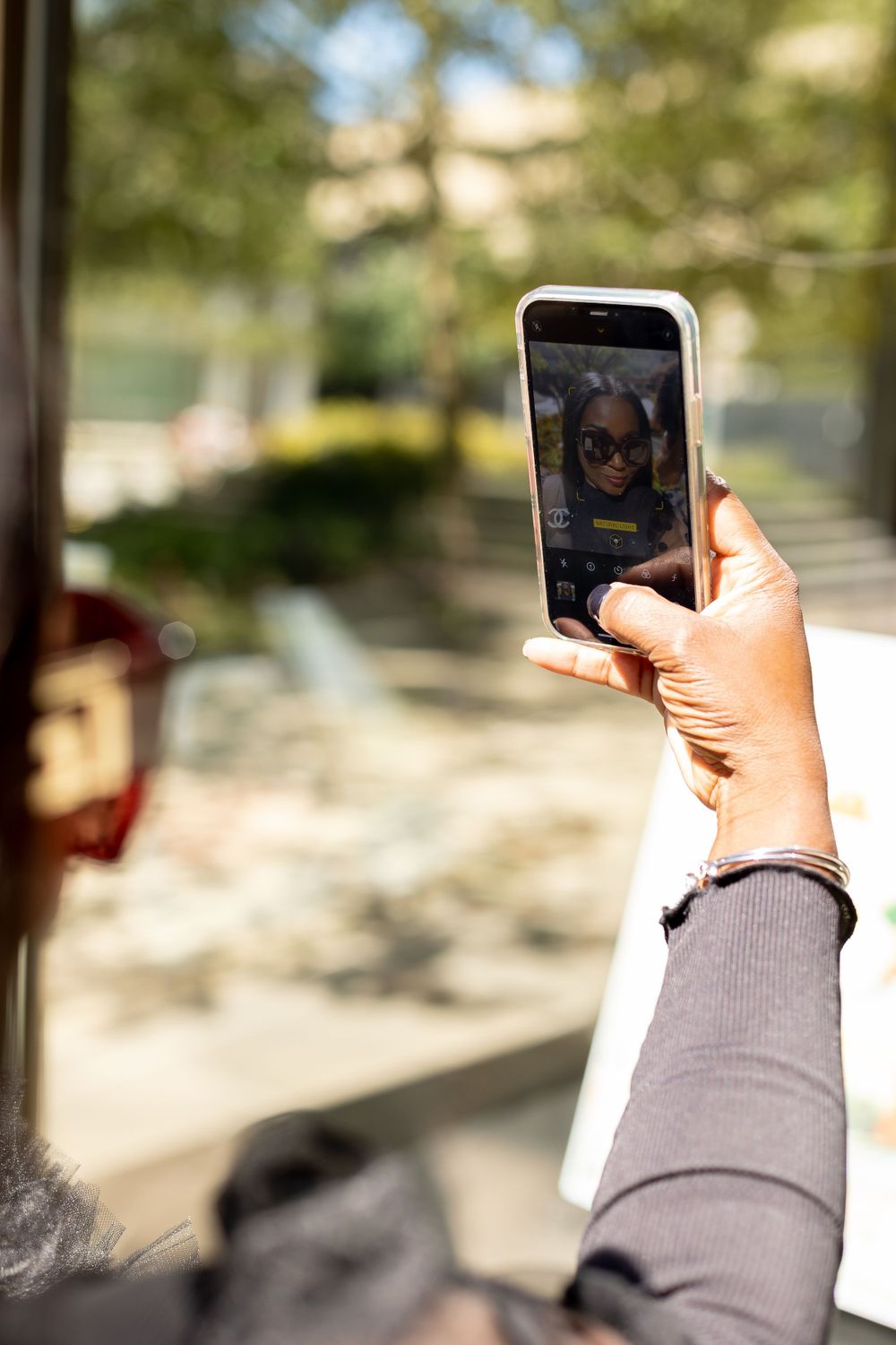 Hand holding a smartphone displaying a selfie of a woman wearing sunglasses outdoors.