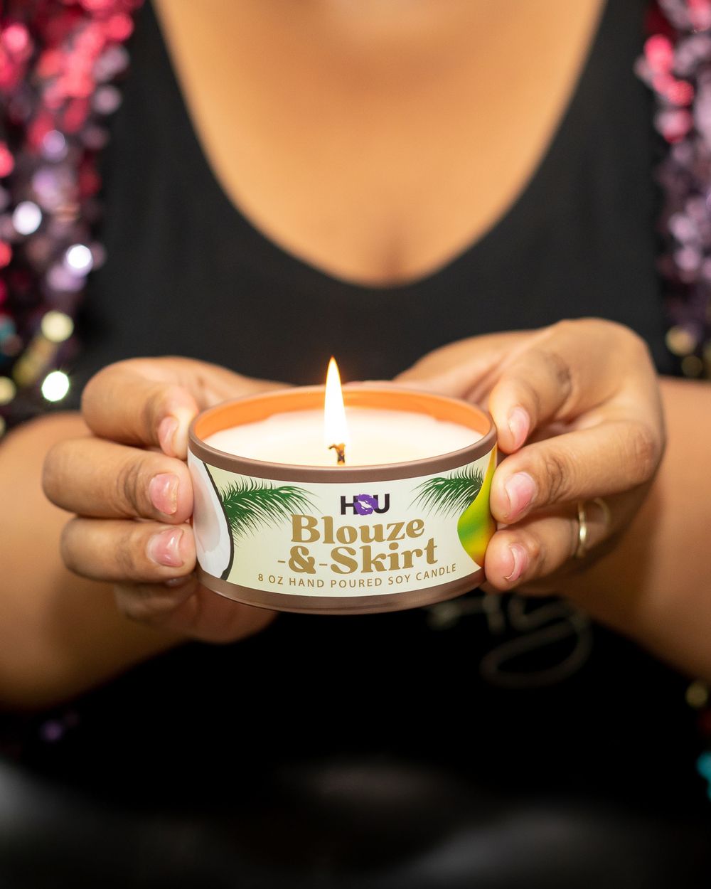 Hands holding a lit HSU 'Blouze & Skirt' hand-poured soy candle in a tin against a sparkly background.
