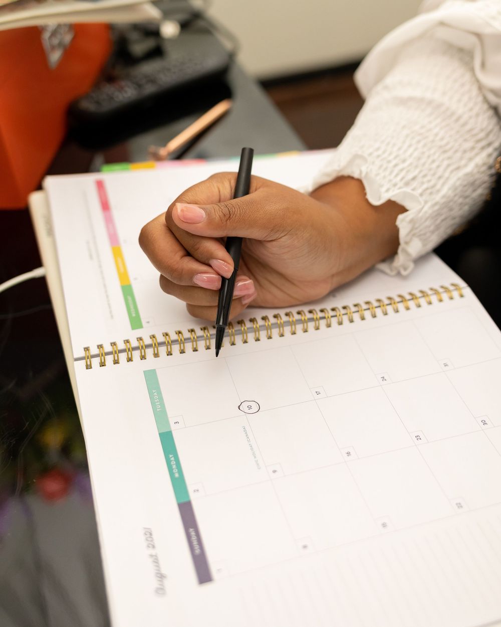 Close-up of a hand writing in a spiral-bound planner with colorful tabs at a desk.