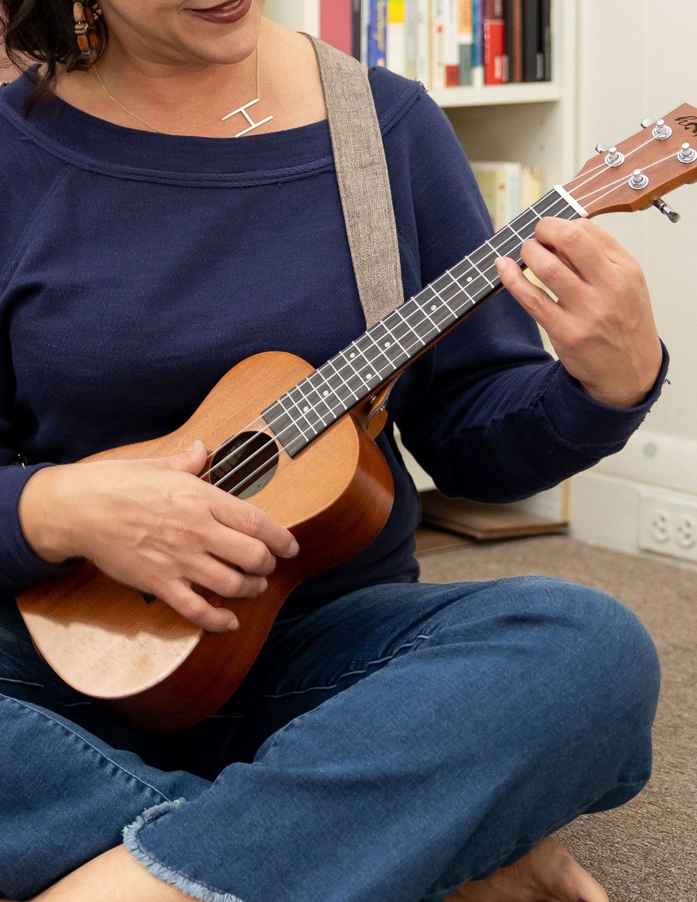 Woman in navy sweater playing a wooden ukulele while sitting cross-legged on the floor.