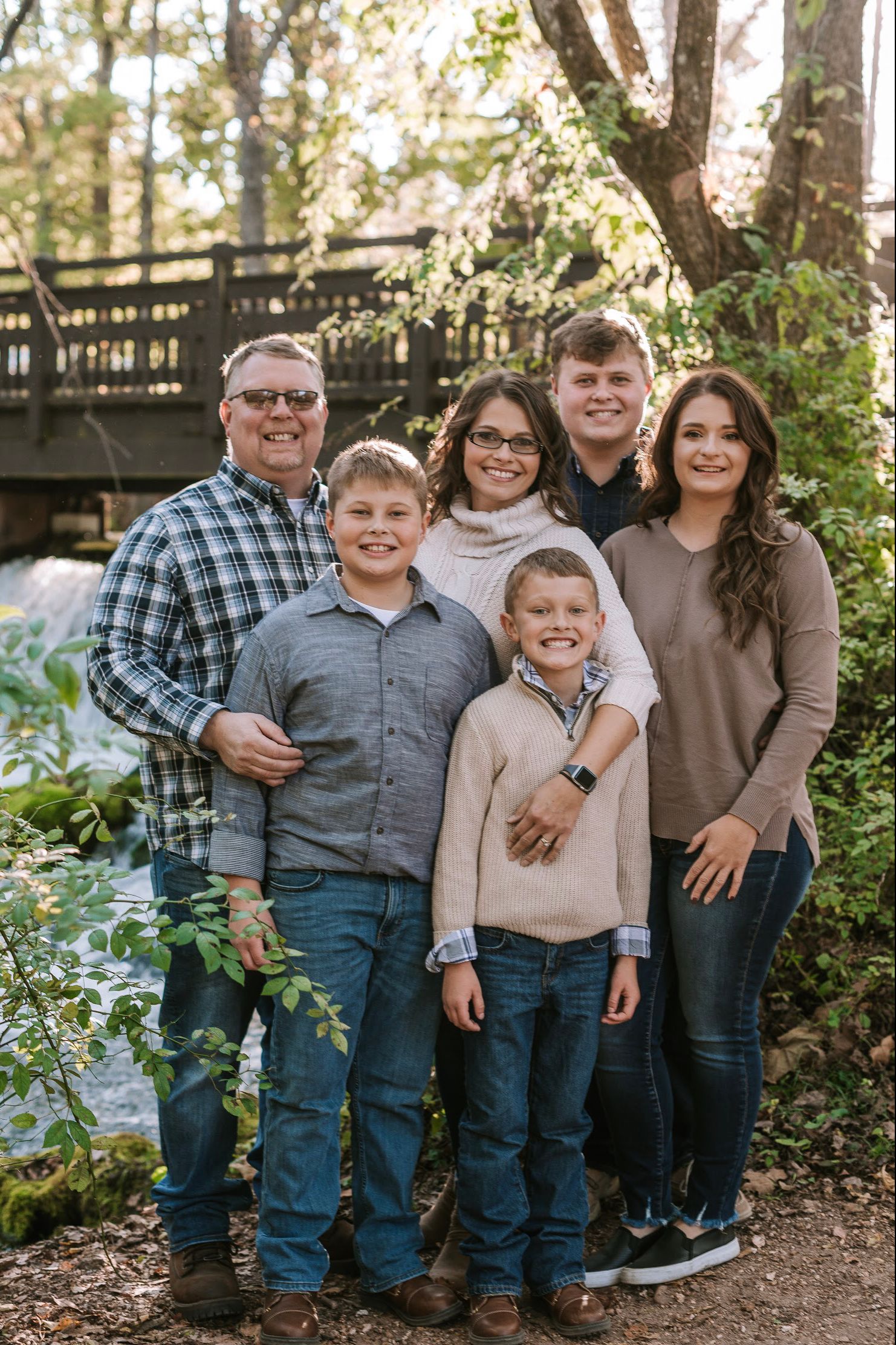 Family portrait of group in casual clothing standing together outdoors near wooden bridge.