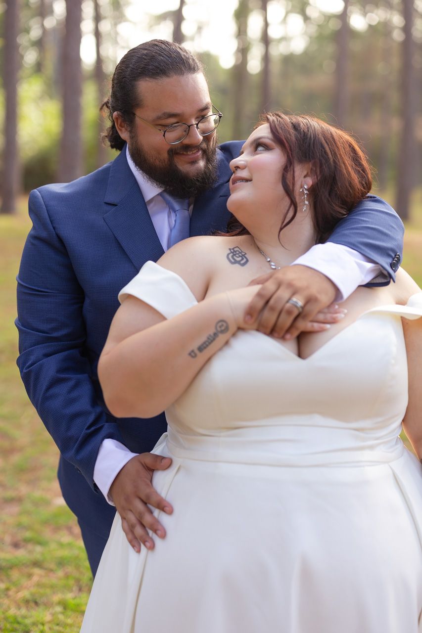 Wedding couple embracing in white dress and navy suit among pine trees.