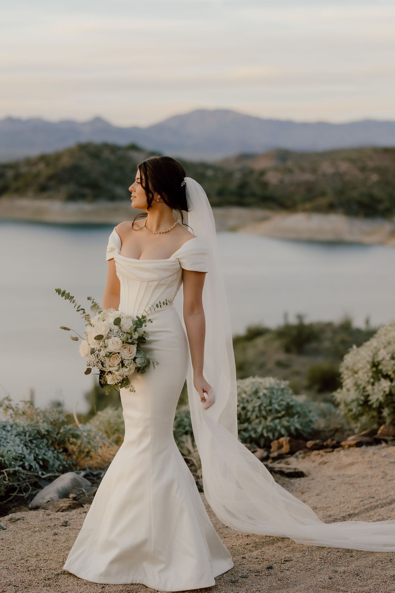 Bride in elegant white mermaid dress poses with bouquet by scenic mountain lake.