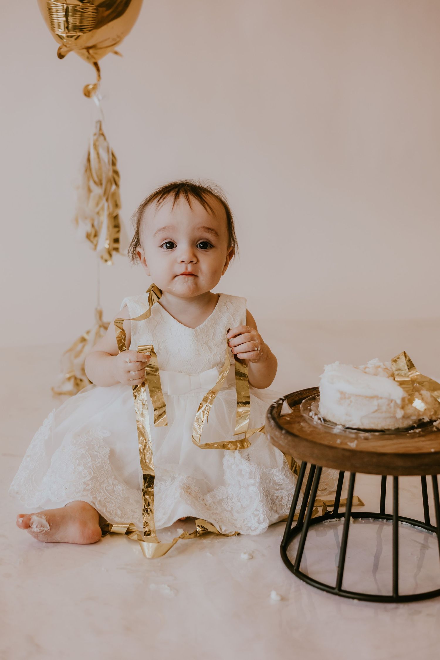 A small toddler in a white lace dress sits on the floor with gold decorations and a cake on a side table.