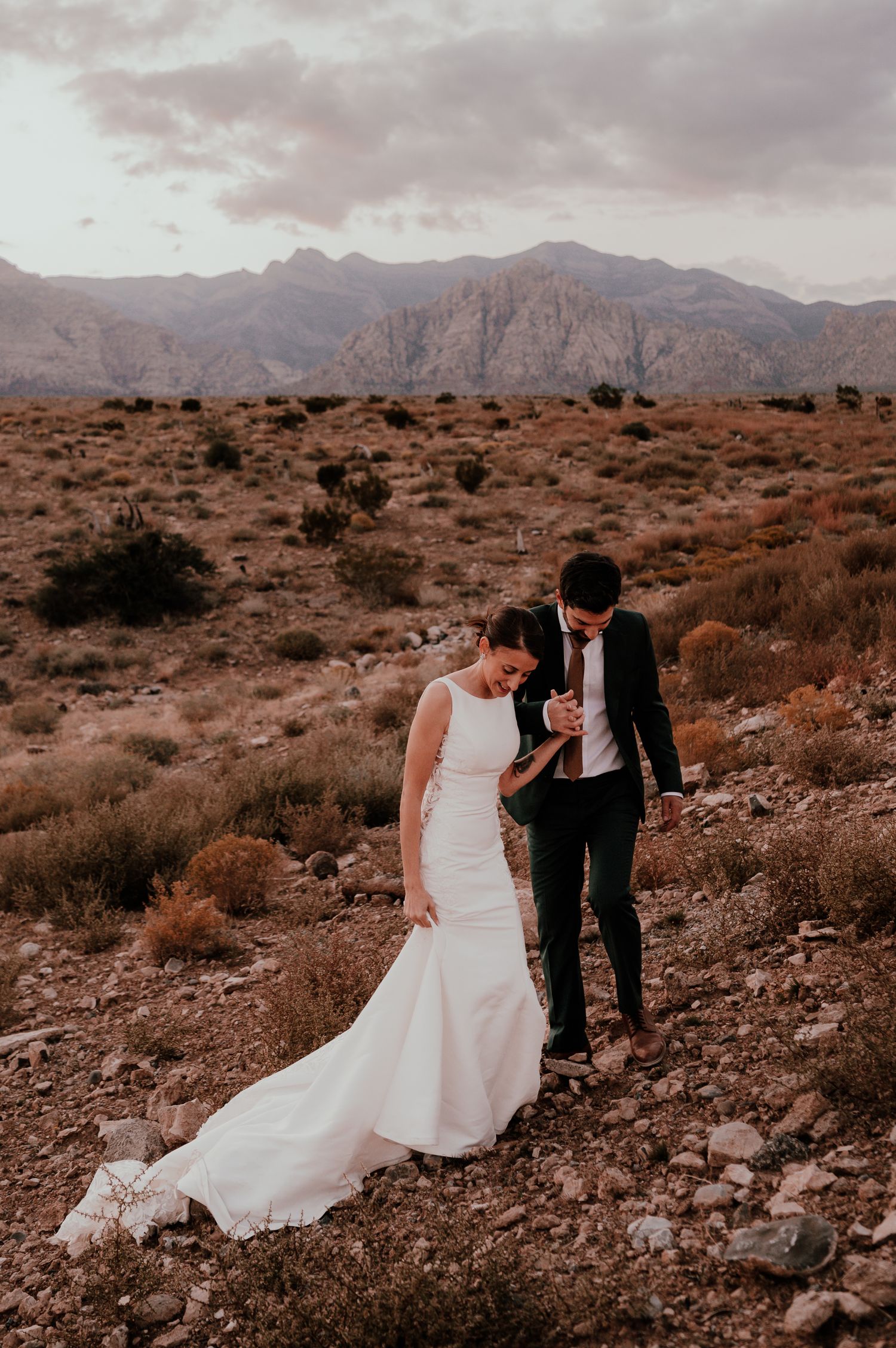 A couple in wedding attire walk together on rocky desert terrain with dramatic mountains and cloudy skies in the background.