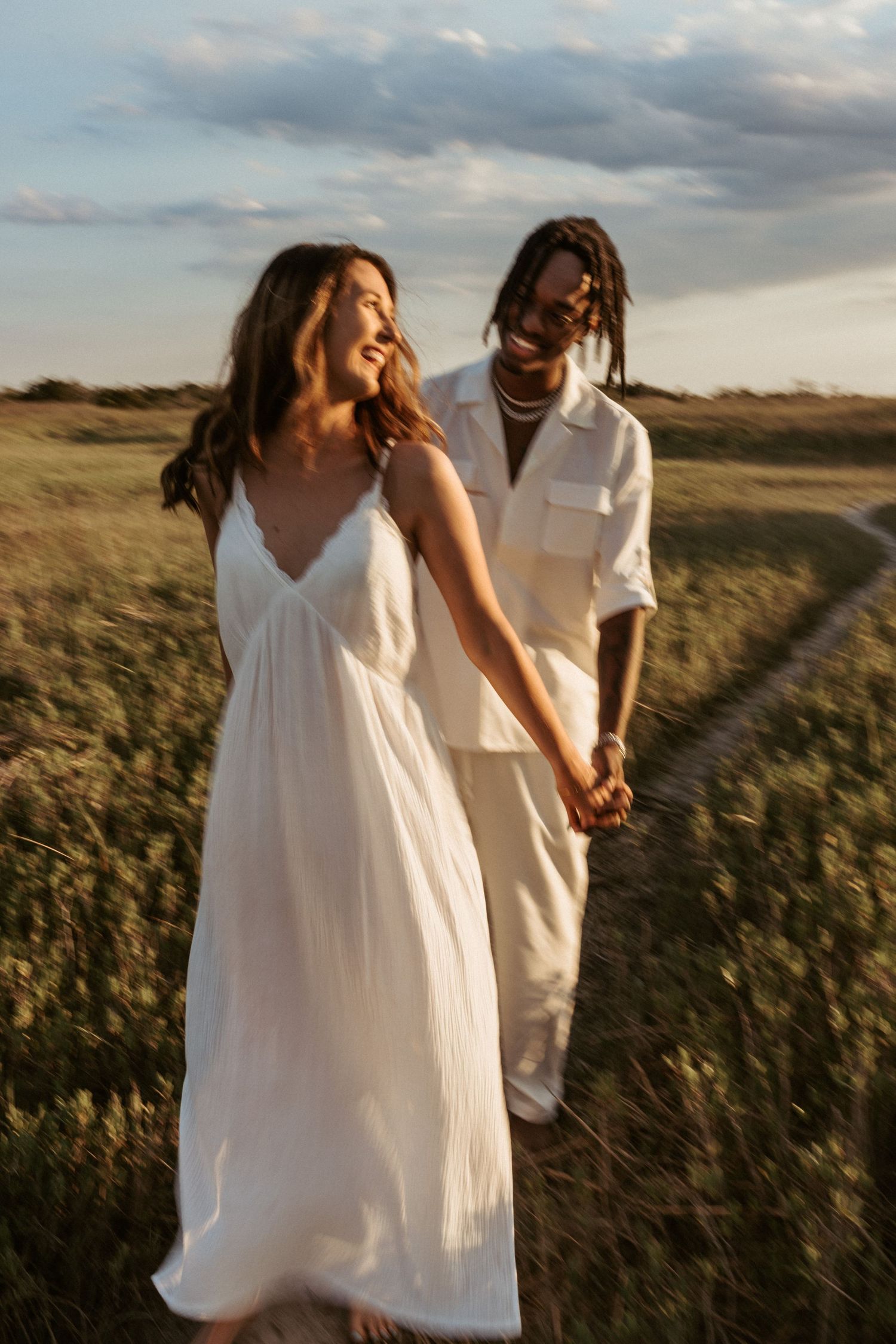 Couple in white outfits walking through a sun-lit field during golden hour.