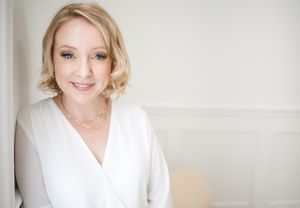 Smiling professional with light curls poses against neutral studio wall, projecting polished and approachable branding energy.