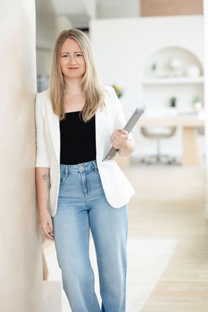 Strathroy therapist leaning against a wall wearing a white blazer, black tank, and blue jeans holding a laptop in hand with a subtle smile