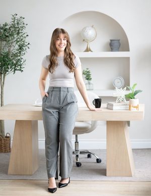 business coach entrepreneur wearing dress pants and a t-shirt learning against a desk with a light coloured background and shelving