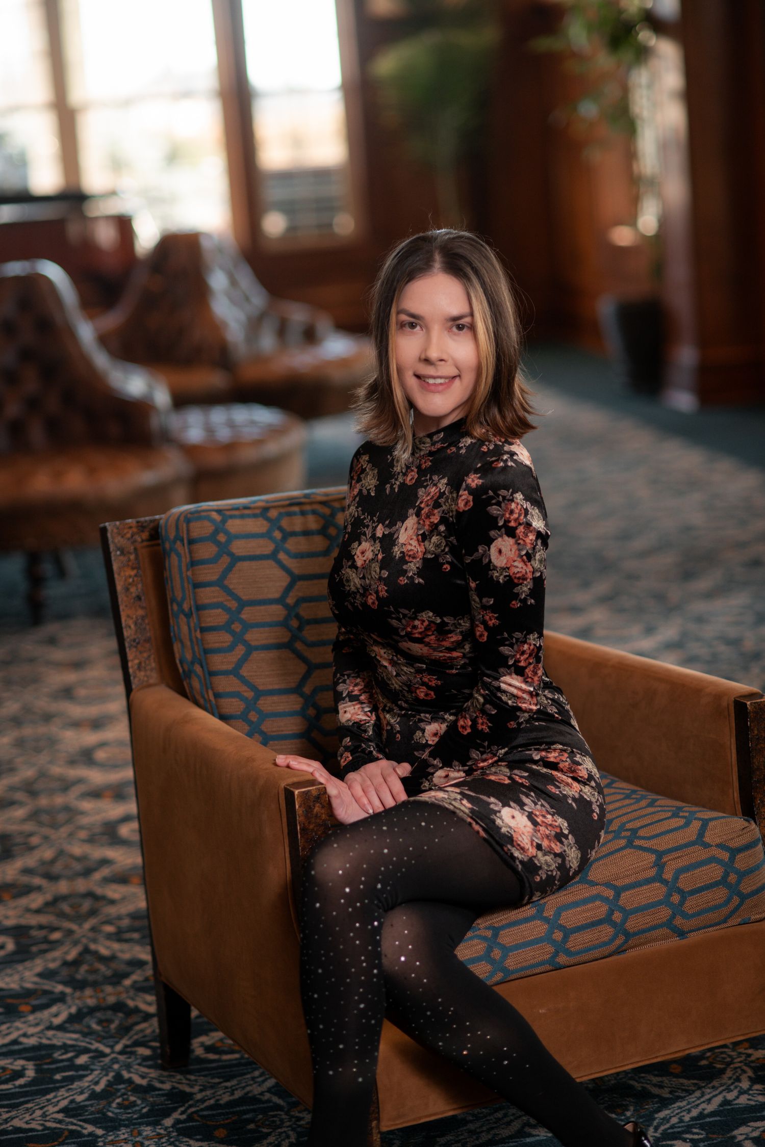 Smiling woman in floral velvet dress sits in armchair in luxurious hotel lounge interior.
