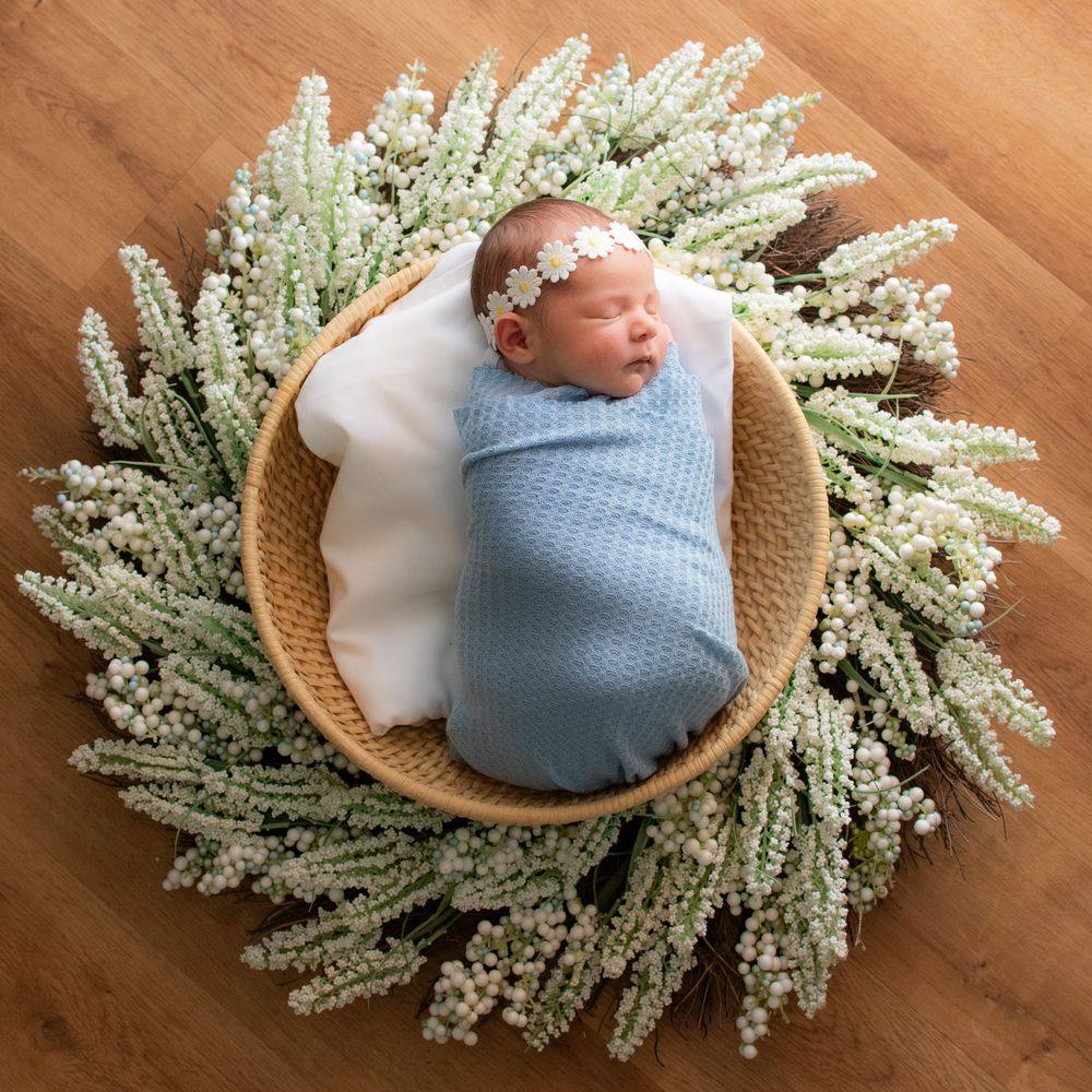 newborn wrapped in blue with daisy headband in a basket on wood floor