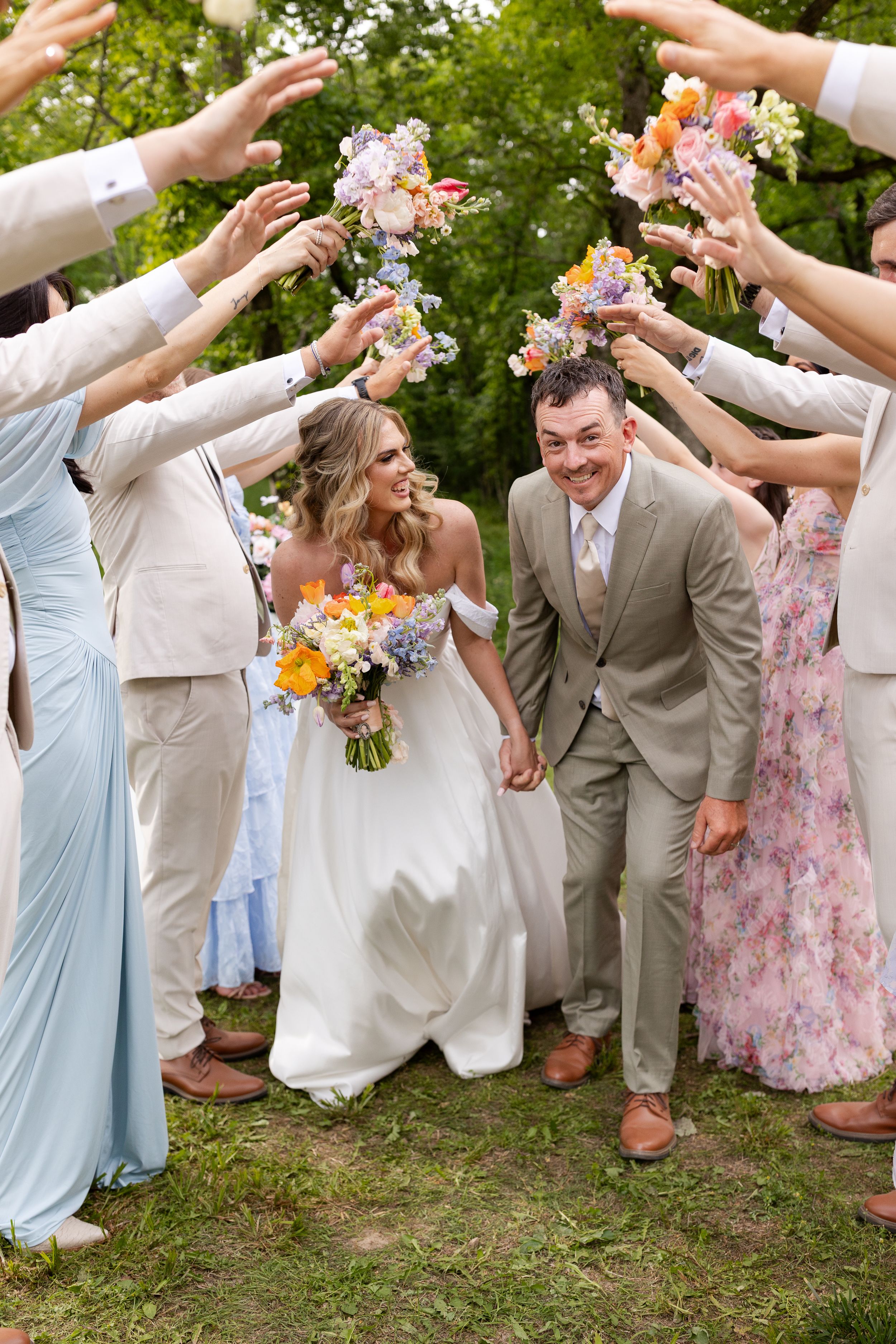 Wedding party guests shower flower petals on couple walking beneath floral arch while holding colorful bouquet.