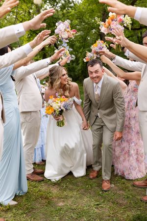 Wedding party guests shower flower petals on couple walking beneath floral arch while holding colorful bouquet.
