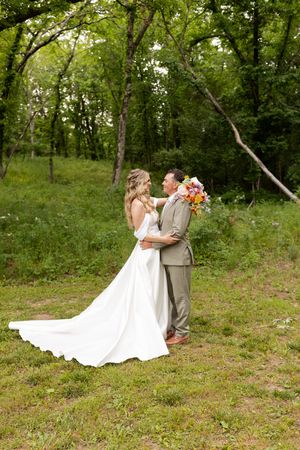 A newlywed couple in a rustic outdoor forest setting, with a flowing white wedding dress and grey suit.