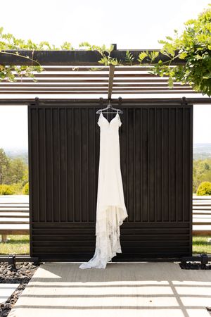 A white wedding dress hangs elegantly against a dark wall with natural lighting and scenic outdoor views.