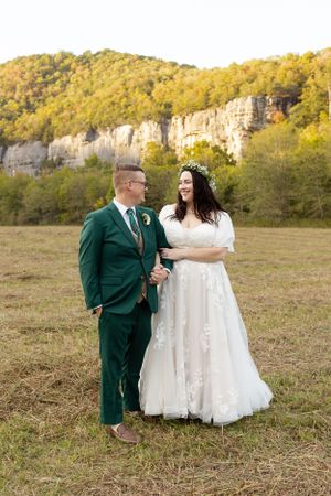 Newlyweds in emerald suit and white gown stand together in mountain meadow.