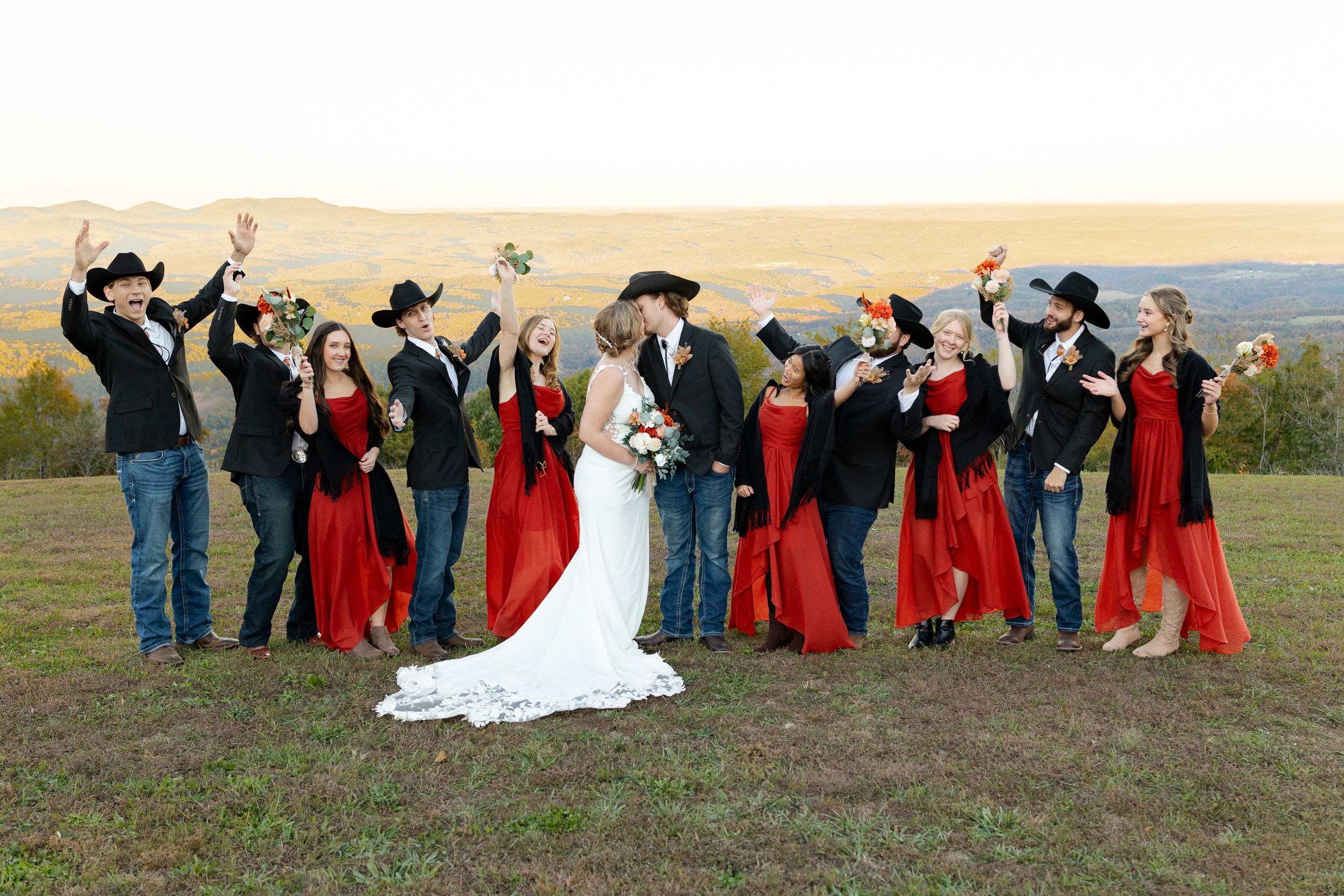 Wedding party in red dresses and cowboy boots pose together in a rustic outdoor setting at sunset.
