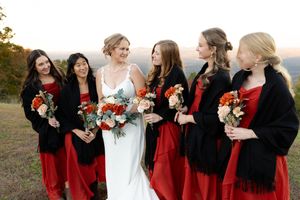 Bridesmaids in red dresses and black shawls laugh together while holding white and red bouquets outdoors.