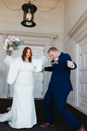 A joyful wedding couple in white dress and navy suit dancing together under a vintage pendant light.
