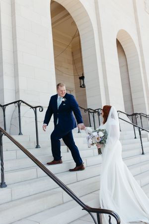 A couple descends white stone steps with ornate railings outside a classic building.