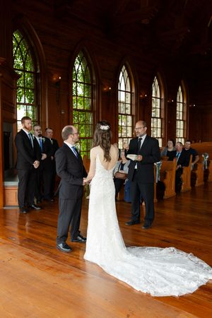 A wedding ceremony takes place in a beautiful chapel with tall arched stained glass windows and hardwood floors.