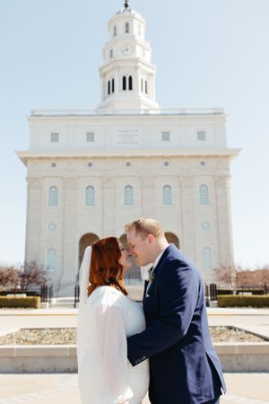 A wedding couple shares an intimate moment in front of a grand white temple building.