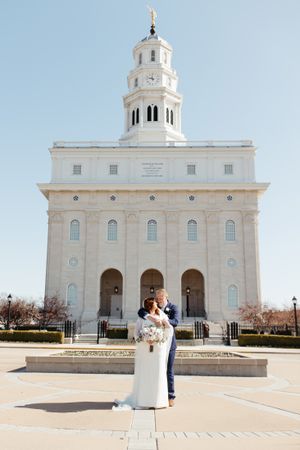 A couple poses for a wedding photo in front of a majestic white temple with a tall spire.