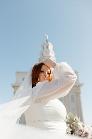 A bride in a long-sleeved white gown poses elegantly against a white church steeple.